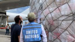 Une troisième cargaison de fournitures essentielles destinées à sauver des vies arrive au Venezuela. Deux hommes, dont l'un porte un gilet bleu des Nations Unies, se tiennent debout devant la cargaison, sur le tarmac de l'aéroport.