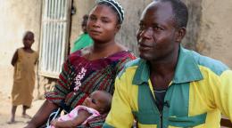 Safiatou et son mari Idrissou sont assis côte à côte. Safiatou tient son nouveau-né dans les bras et le couple coupe explique comment il a pu éviter les démarches fastidieuses liées à la déclaration de la naissance de leur cinquième enfant grâce au nouveau service téléphonique mis à disposition gratuitement.