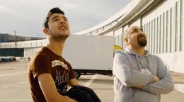Deux hommes se tiennent debout sur le tarmac d’un aéroport, devant un gigantesque bâtiment, les yeux rivés vers le ciel.