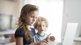 تظهر فتاة صغيرة تستخدم جهاز كمبيوتر محمول، بينما يقف شقيقها الأصغر مندهشاً.