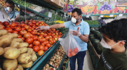 Abdou, un réfugié yéménite à Amman, fait ses courses chez un marchand de légumes après la réouverture des petits commerces. Il porte un masque de protection et des gants. Au premier plan de l’image, un enfant, lui aussi portant un masque, le regarde mettre des tomates un sac en plastique.