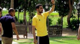Mohamed ElKholy está parado en el centro de la imagen, en el fondo se aprecia un parque con grama. Mohamed está usando un pantalón de chándal negro y una camiseta amarilla. Levanta el brazo izquierdo para llamar la atención a otra persona.