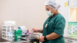 Une infirmière portant une charlotte et un masque chirurgical prépare du matériel destiné à la réalisation d’une prise de sang.