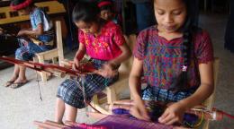 Photo showing three indigenous girls sitting on a chair and learning to weave using a traditional loom 