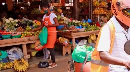 Un conducteur de SafeBoda quitte le marché à vélomoteur pour aller livrer de la nourriture. Il porte un masque et un casque de protection. En arrière-plan, une femme remplit un sac de fruits sur un des nombreux étals de fruits et légumes du marché.