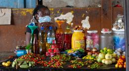 Une femme portant un masque de protection est assise derrière un étal du marché de Freetown. Sur son étal se trouvent toutes sortes de denrées alimentaires fraîches et en boîte.