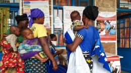 تقف نساء مع أطفالهن خارج مرفق طبي ويقرءن لافتات حول فيروس كورونا والتطعيمات.