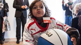 Un niño vestido con un chándal deportivo, sostiene una pelota de baloncesto con ambos brazos y mira a la cámara, haciendo el gesto de estar mandando un beso, con los labios fruncidos.