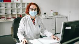 Snežana Bursać Aranđelović está sentada en su escritorio con una computadora (frente a ella) en el Instituto de Salud Pública del Cantón de Sarajevo. Lleva una bata blanca y una mascarilla quirúrgica azul y está mirando a la cámara. Podemos ver detrás archivos y mobiliario de oficina colocado en estanterías blancas.