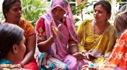 Un grupo de mujeres indias, vestidas con ropas tradicionales de colores brillantes, se sienta en el suelo y conversa.