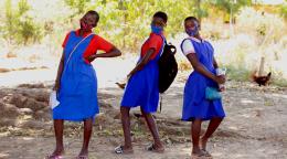 Tres colegialas con mascarillas protectoras se divierten posando para la foto, en el patio de la escuela primaria de Luwambaza. Las tres tienen el pelo corto y visten uniformes escolares, atuendos de color azul brillante.