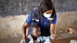 En Zimbabwe, en el aula de una escuela local, una miembro del personal de la OMS que lleva una mascarilla protectora ayuda a un niño pequeño a extender la mano y agarrar, por sí mismo, un medicamento. Una mano de un adulto la entrega la medicación, en formato de pastilla, con una cuchara. Al fondo, vemos una pizarra.