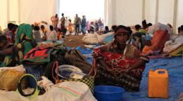 Une femme éthiopienne réfugiée est assise, face caméra, sous une immense tente, sur une grande bâche bleue en plastique. Elle est entourée d’objets divers, dont un couffin, un bidon orange, de la vaisselle, une couverture, une bassine bleue. Derrière elle, en arrière-plan, on aperçoit de nombreux autres réfugiés, hommes femmes et enfants.