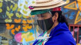 Gros plan sur une femme autochtone se tenant devant une peinture murale haute en couleurs colorée. La femme porte un masque noir et une visière de protection et est vêtue d’un habit à dominante bleue ainsi que d’un chapeau traditionnel.