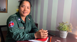 L'officière de police Kong Ravin est assise à une table où sont posés un cahier, un stylo, un téléphone portable et un petit pot de fleurs. Elle porte un uniforme et sourit à la caméra.