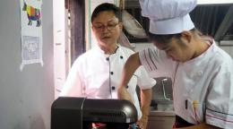 A man oversees a student chef as she stirs ingredients in a mixer.