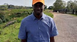 Un homme sourit avec fierté à la caméra. Il portant une casquette orange qui témoigne de son soutien à la lutte contre la violence de genre. Il se tient sur une route bétonnée traversant un champ verdoyant.