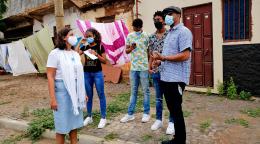 La Coordinadora Residente, Ana Patricia Graça, habla con jóvenes artistas en un semi círculo, frente a las puertas de las casas de una comunidad. Todos usan mascarillas.