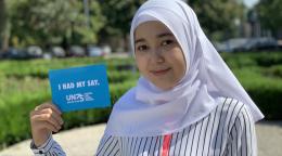 Une adolescente portant un foulard sourit discrètement à la caméra en tenant une petite carte bleue sur laquelle on peut lire "I had my say" et "UN75", qui signifient "J’ai pu exprimer mes idées" et "ONU75".