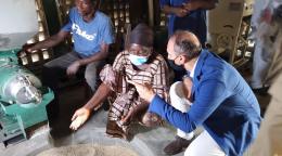 Resident Coordinator, Vincent Martin kneels by a woman community leader as they speak near a machine that is filling a round hole with grains.