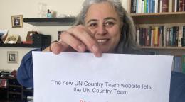 A woman smiles cheerfully at the camera as she holds a sign that reads "The new UN Country Team websites lets the UN Country Team BE MORE VISIBLE"
