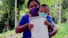 A mother wearing a protective face mask gazes to the left of the camera as she holds her son, also wearing a face covering. He proudly holds up his registration.