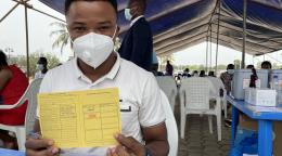 A man in a white shirt and white face mask holds up a yellow COVID-19 vaccination card. 