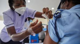 A nurse in a purple shirt and white face mask administers a covid vaccine on a woman with a blue shirt and cap.