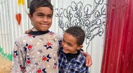 Two small boys with arms around each other smile happily as they stand outside by a wall that has a mural painting.