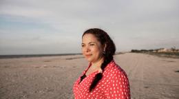 Woman in a red shirt looks out into the distance at a sandy location. 