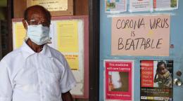 A man with a white shirt and a white face mask stands in front of a bulletin board with information on COVID-19. 