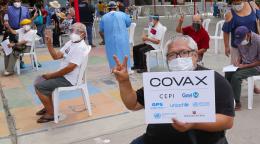 Una persona sostiene un letrero de COVAX mientras varias otras personas levantan los pulgares y hacen signos de paz a la cámara.