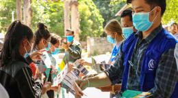 Plusieurs membres du personnel de l'OIM portant des vestes bleues et des masques de protection distribuent des brochures d'information à un groupe de jeunes femmes qui portent elles aussi des masques de protection.