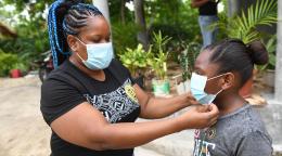 Dans une cour extérieure bordée d’arbustes, une femme ajuste le masque de protection de sa fille, une élève de l'école primaire de Little Bay. Little Bay est une commune principalement composée de pêcheurs située à Westmoreland, à l'extrémité ouest de l'île de la Jamaïque.