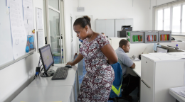 Dans un bureau d’entreprise, une femme vêtue d’une robe travaille, debout, sur un ordinateur, tandis qu'un homme est assis à un bureau, à quelques pas, devant son propre ordinateur.