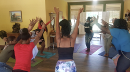 Varias mujeres, en una habitación amarilla, tienen sus manos mientras practican yoga en grupo.
