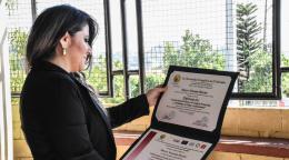 A woman with dark hair and a black jacket looks down at her diploma.