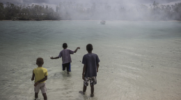 Trois enfants se tiennent debout dans un plan d'eau, dos à la caméra et regardent un bateau s’éloigner.