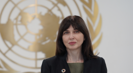 A woman, standing in front of the United Nations symbol, wearing a black jacket and green shirt looks directly at the camera. 