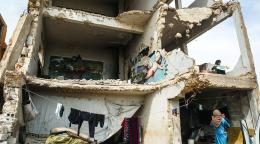 一名身穿蓝色连衣裙的妇女和孩子们站在一栋部分被毁的建筑内和附近。