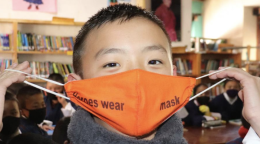 A young boy puts on his orange face mask while looking straight at the camera. 