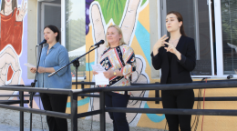 Three women stand in front of a building decorated with many colors. A woman, dressed in black, on the end is translating the speech to sign language.