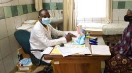Un médecin en blouse blanche est assis à son bureau, dans un cabinet de consultation, le regard tourné vers la caméra, en face d'une patiente. Tous deux portent un masque de protection.