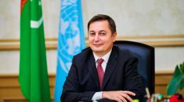 رجل يرتدي بدلة يبتسم للكاميرا وخلفه علما تركمانستان والأمم المتحدة.