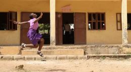 一个身穿粉色衬衫和紫色连衣裙的年轻女孩在一栋教学楼附近蹦蹦跳跳。