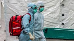 Two health care workers in full body medical gear and face masks walk with a large red backpack. 