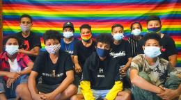 Un grupo de personas jóvenes con máscaras se sientan con las piernas cruzadas delante de una bandera con los colores del arco iris.