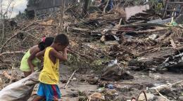 两个孩子手里拿着一个袋子，在一栋房屋的废墟附近行走。