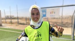 Sur un terrain de football, une jeune fille tient un ballon de football sous le bras et lève le pouce de l'autre main en souriant à l'objectif. Elle porte un foulard blanc sur la tête et est vêtue d’un maillot de couleur jaune portant le logo de l’UNICEF et celui du programme Makani.