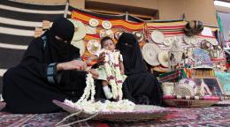 A Saudi woman sits on a rug and threads fresh flowers to sell in a souk as another woman sitting beside her holds up a baby. 